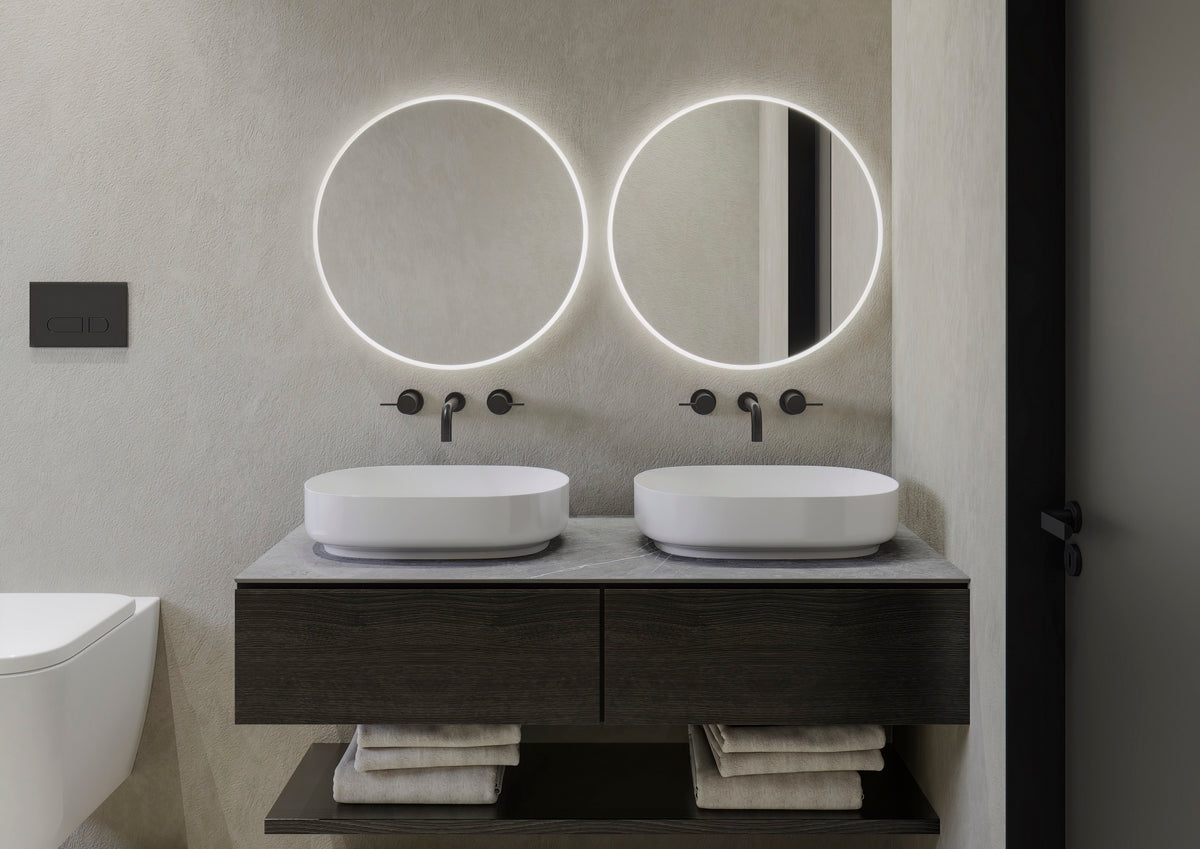 Dos espejos CLARK redondos encendidos, con aro de luz frontal y halo ambiental, sobre un doble lavabo con mueble de madera oscura (foto del fabricante)
