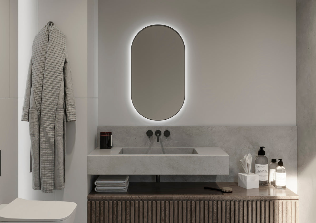 Espejo de baño en forma de cápsula con marco negro y luz retroiluminada encendida sobre un lavabo de piedra, modelo MILUS